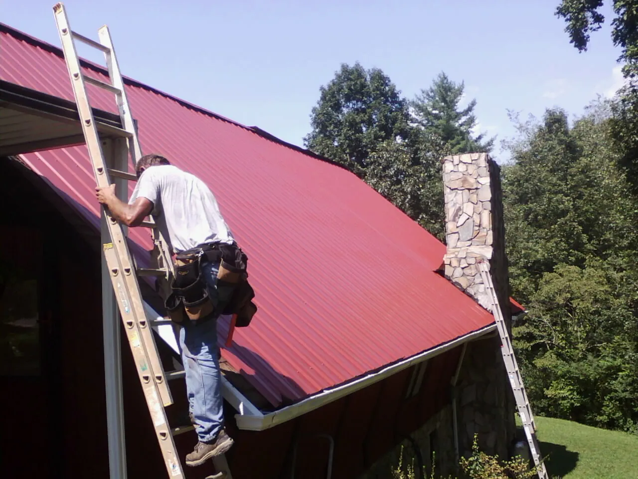 Roofer on a ladder beside a red metal roof and stone chimney — Soffit & Fascia Repair by Roofing Brothers in Rangely